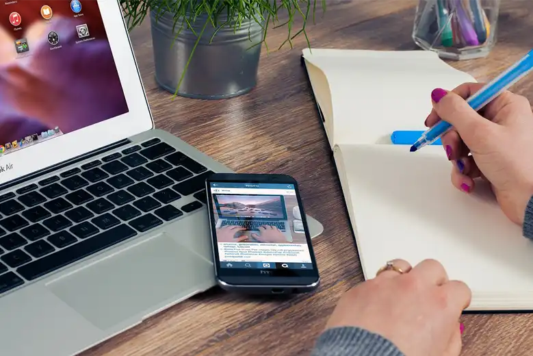 Mujer escribiendo en bloq de notas junto a su laptop y teléfono celular